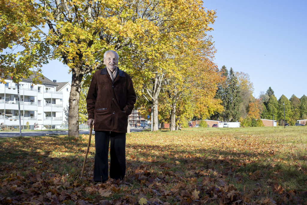 Eldre mann med stokk står i en park foran et stort tre.
