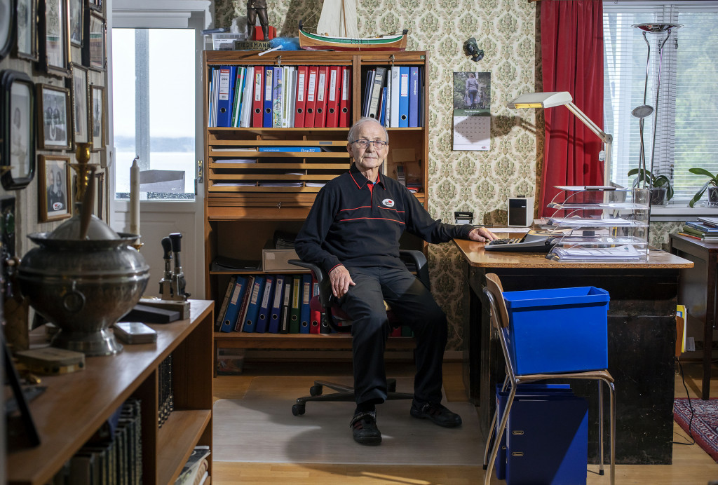 Eldre mann sitter på et kontor som har fullt av permer og familiebilder.