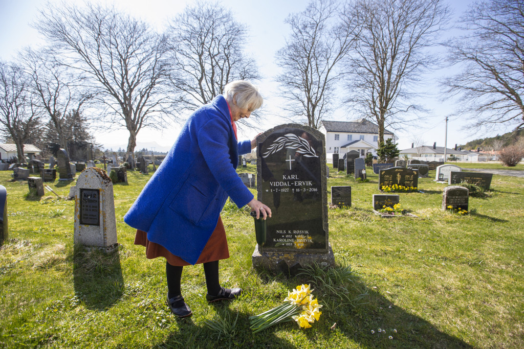 Dame i blå jakke står ved en gravstøtte på kirkegården.